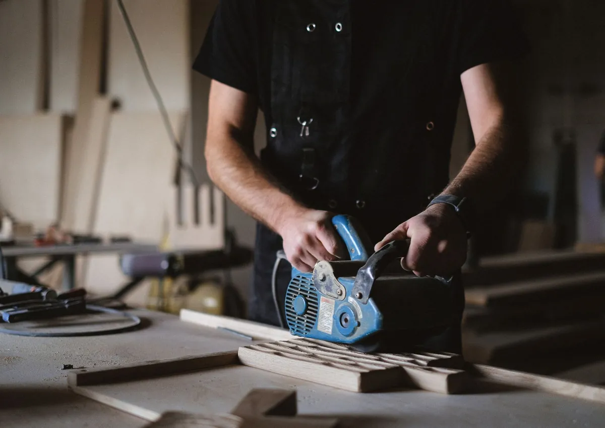 Unrecognizable male carpenter using belt sander while working with wooden plank at workbench in joinery with professional equipment on blurred background